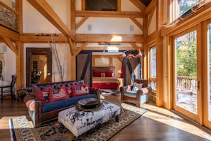 Un salon spacieux avec un grand canapé, une table basse et une cheminée. Le plafond est en bois, avec des poutres apparentes, et de grandes fenêtres offrent une vue sur l’extérieur.