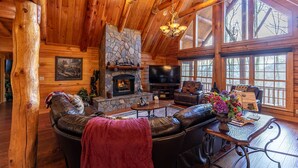 A log cabin interior with a stone fireplace, leather furniture, and large windows. - Running Waters Lodge - private log home with hot tub, game room, fire pit! (Banner Elk)