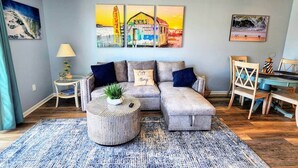 A living room with a sectional sofa, a coffee table, and a dining area with a table and chairs. There are wall art pieces above the sofa and a painting on the wall to the right.