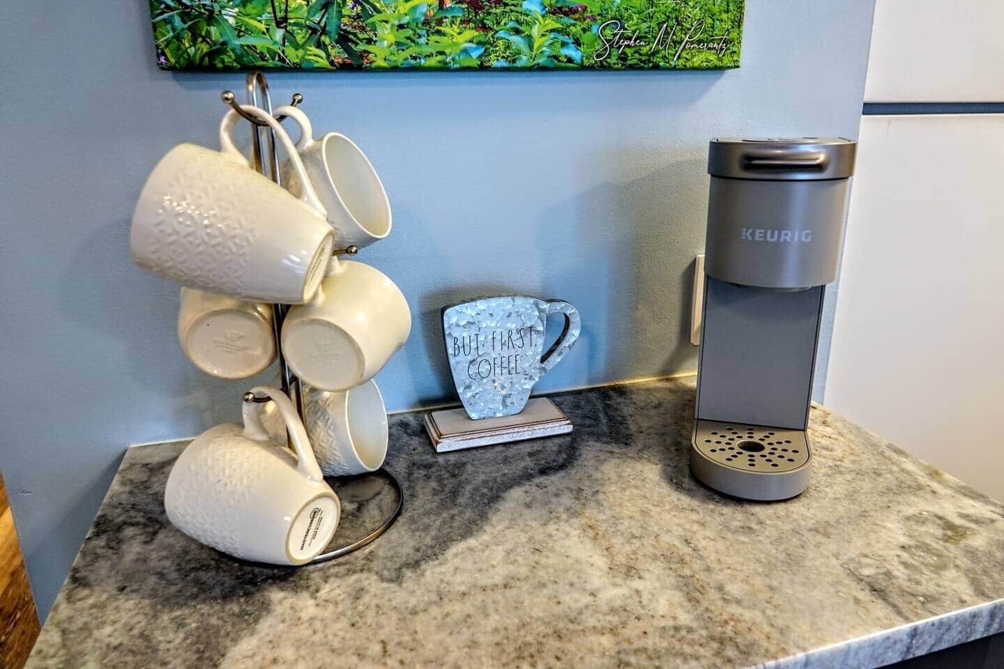 Een keukenblad met een Keurig koffiezetapparaat, een stapel witte mokken en een decoratieve mok waarop 'BUT FIRST COFFEE' staat.