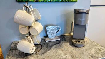 Een keukenblad met een Keurig koffiezetapparaat, een stapel witte mokken en een decoratieve mok waarop 'BUT FIRST COFFEE' staat.