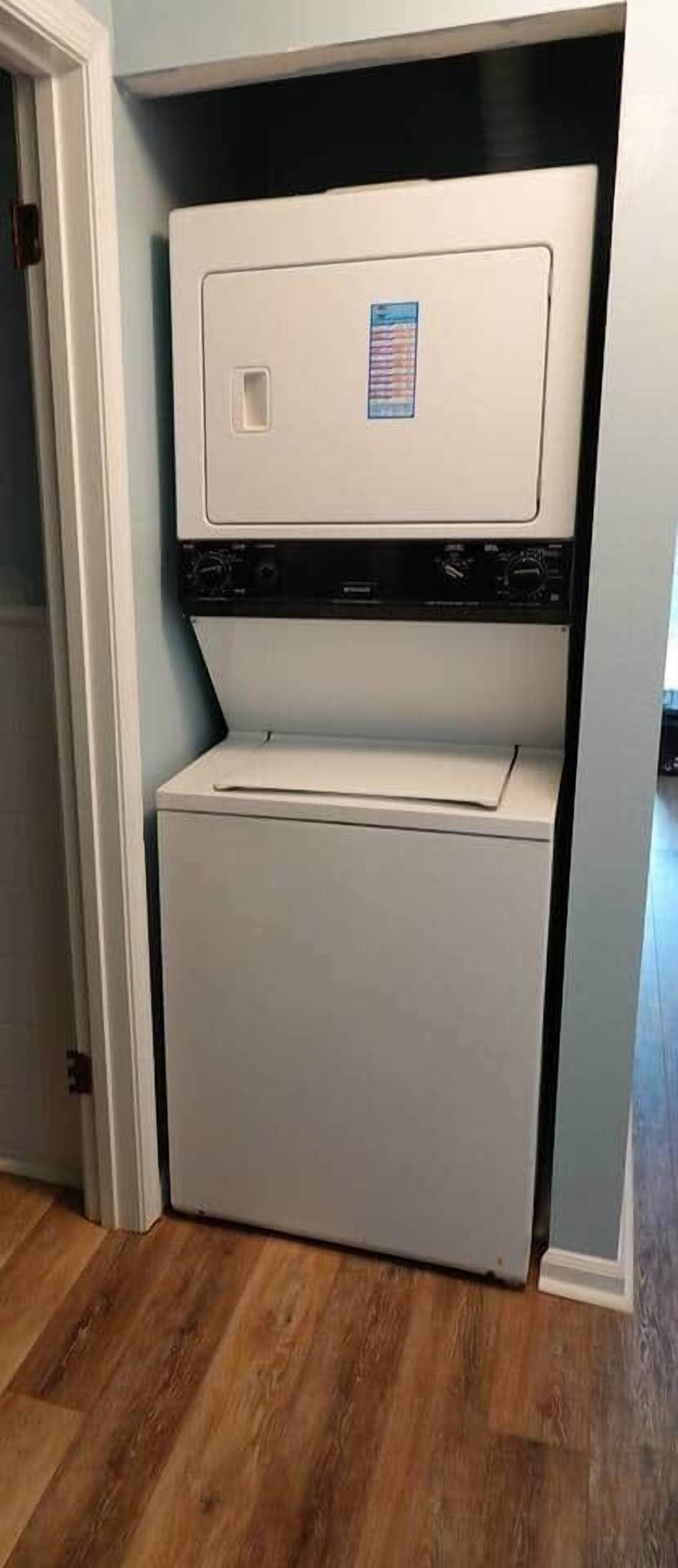 A compact washer and dryer unit in a corner of a room with wooden flooring.