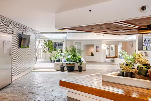 Un lobby moderno de hotel con un muro de diseño geométrico, plantas en macetas, un banco de madera y suelo de baldosas.