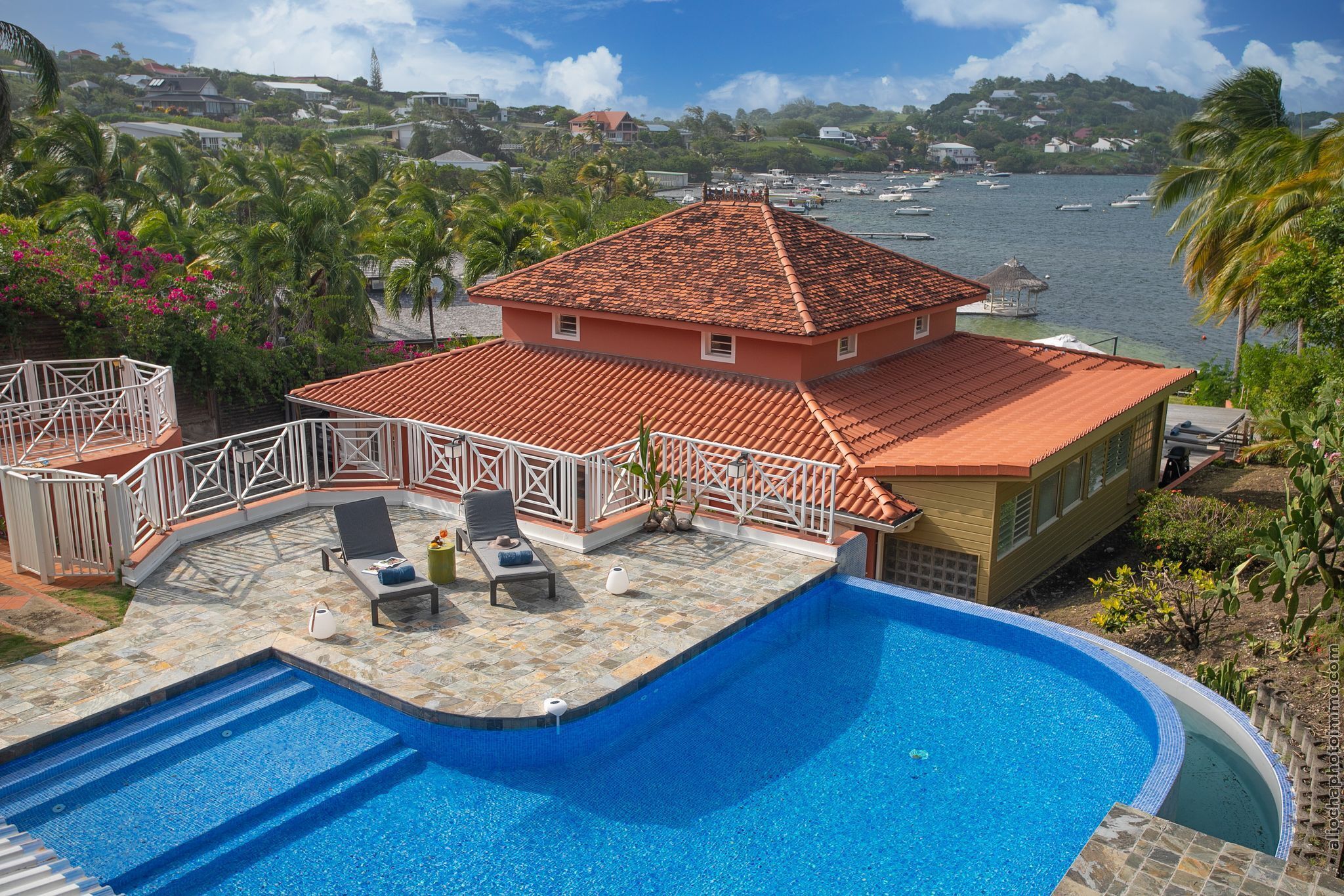 A swimming pool with a tiled deck, lounge chairs, and a view of a body of water with boats.