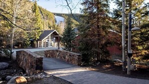 Un ponte di pietra conduce a un lodge con rivestimento in legno, immerso tra alberi e con montagne sullo sfondo.