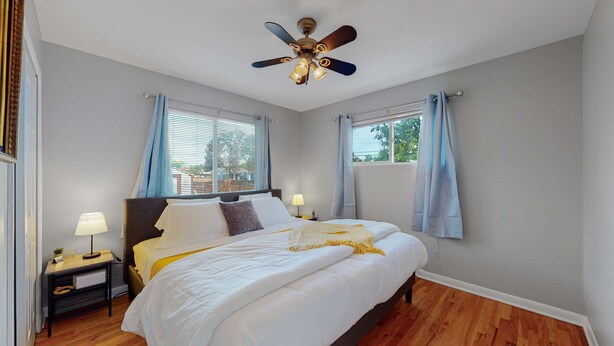 Un dormitorio con cama, mesitas de noche, ventilador de techo y ventanas con cortinas.
