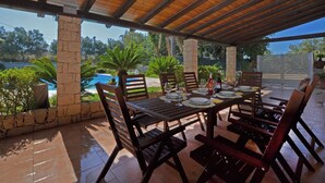 En udendørs terrasse med et træbord dækket til spisning, omgivet af stole, med en pool og grønt i baggrunden.