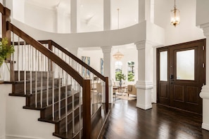 A grand staircase leading to a high-ceilinged room with a chandelier, a wooden door, and a view of the outdoors.