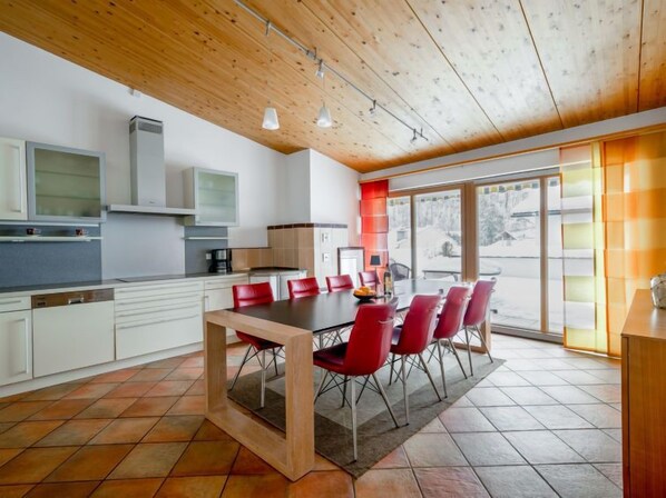 A modern kitchen with white cabinets, a wooden ceiling, and a dining area with red chairs and a glass table.