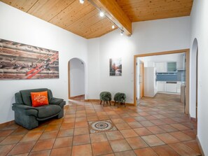 A spacious living area with a wooden ceiling, a sofa with a 'Welcome home' pillow, a patterned rug, and a view into a kitchen through an open doorway.