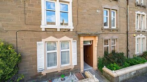 A two-story brick building with white-framed windows, a small entrance door, and a visible garden area with potted plants and gravel. - Manor Place - Convenience and Style (Broughty Ferry, Dundee)