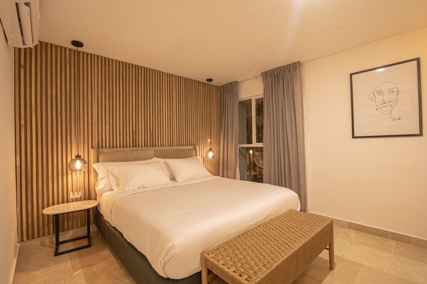 A modern hotel room with a large bed, a small table, a bench, a wall-mounted lamp, and a framed picture.