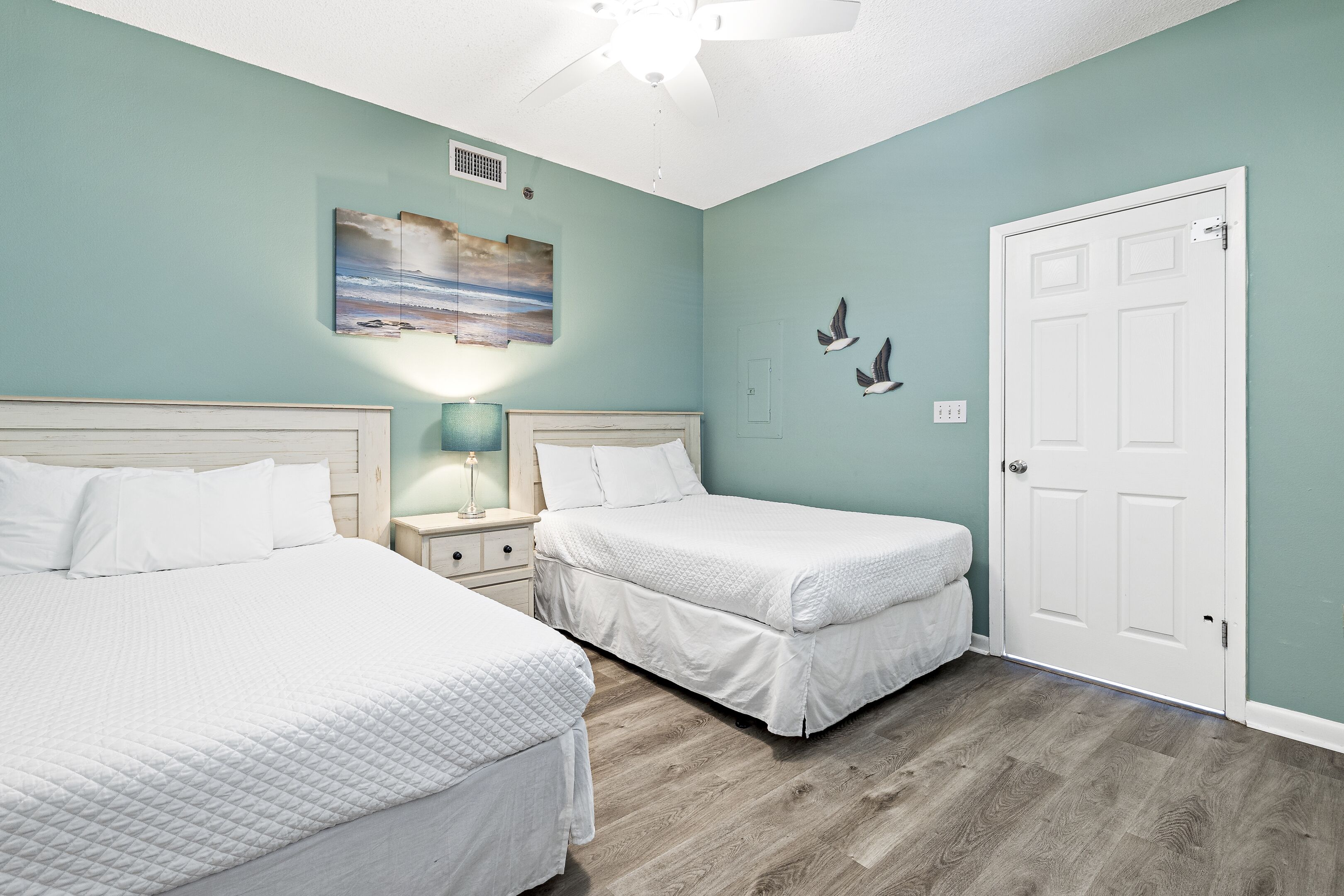 Une chambre avec deux lits, un ventilateur de plafond, un tableau et une porte blanche.