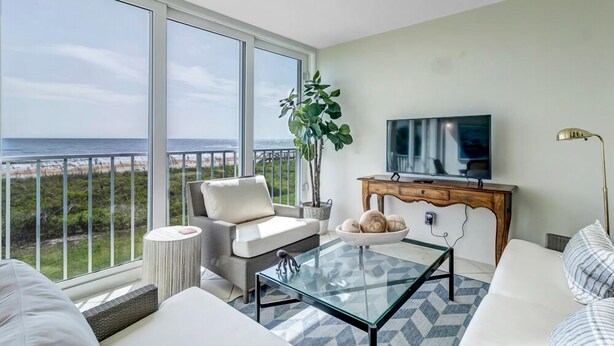 A modern living room with a large window overlooking the beach, a comfortable sofa, a glass coffee table, and a television.