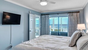 A bedroom with a bed, a ceiling fan, a TV mounted on the wall, and a view of the outdoors through a door.