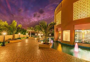 Vista nocturna de un hotel con piscina, palmeras y detalles arquitectónicos iluminados.