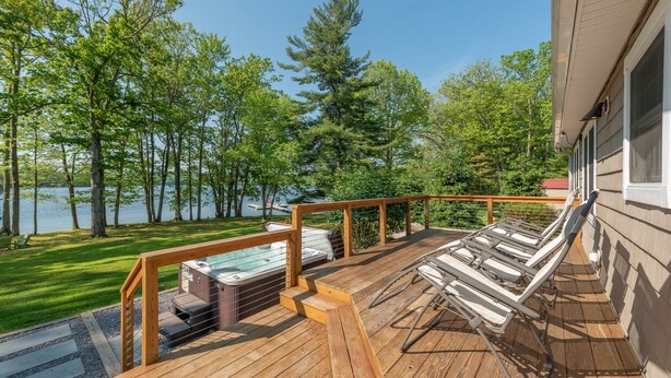 A wooden deck with a hot tub, lounge chairs, and a view of a lake surrounded by trees.