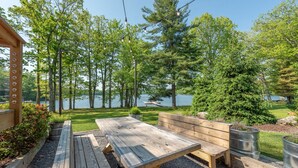 An outdoor seating area with wooden benches and tables, surrounded by trees and a lake view.