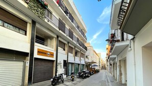 Une rue étroite bordée d’immeubles à plusieurs étages, dont certains ont les volets fermés et d’autres présentent des balcons apparents.