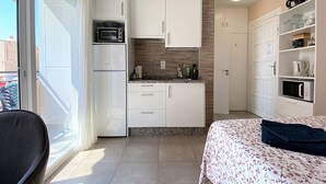A compact hotel room with a kitchenette, a bed with a floral bedspread, a chair, and a view of the outside.