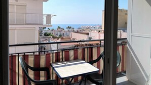 A balcony with a table and chairs, overlooking a coastal cityscape with buildings and the sea.
