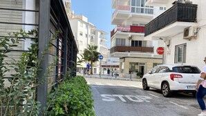 A street with a white car parked on the right, a stop sign, and buildings with balconies and signage.