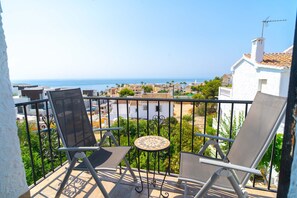 Un balcon avec deux chaises et une petite table, d’où l’on peut admirer une ville côtière avec ses bâtiments et la mer.