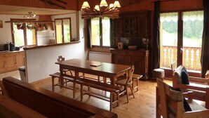 Interior de una cabaña de madera con mesa de comedor, bancos, una zona de cocina y un balcón con vistas.