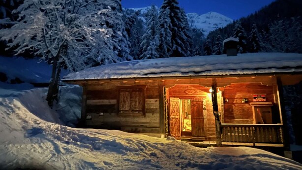 Una cabaña de madera con techo nevado, un porche y un interior iluminado.