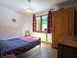 Un lit simple avec une couverture, une armoire en bois, un radiateur, une fenêtre avec des rideaux et un luminaire suspendu.