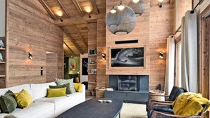Una sala de estar acogedora con un sofá blanco, una mesa de centro oscura y sillones de cuero. Hay una chimenea, paredes de madera y lámparas colgantes esféricas.