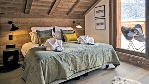 Un dormitorio acogedor con techo de madera, una cama grande con muchas almohadas, una mesita de noche y una ventana con vistas a un paisaje nevado.