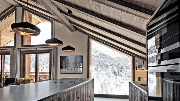 Una cocina moderna con armarios de madera, una isla grande, luces colgantes y vistas a montañas nevadas a través de amplios ventanales.