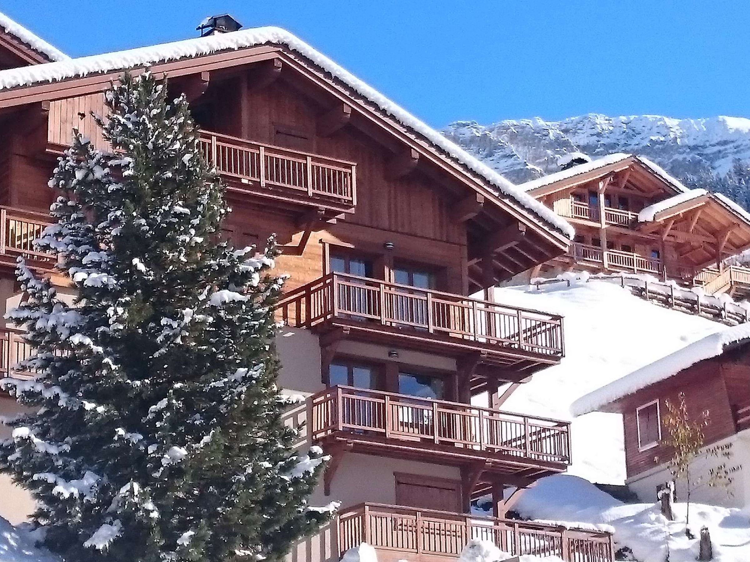 A wooden chalet with balconies, snow-covered roofs, and a mountain backdrop.