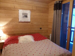 A wooden cabin room with a bed, a framed picture on the wall, and a window with curtains. - Modern Apartment - 2BR - Sleeps 6 - Balcony - Mountain Views - Ski Shuttle - Village Center Nearby (Arêches-Beaufort)