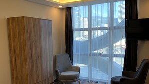 Une chambre d’hôtel comprenant un lit, une armoire, deux fauteuils, une télévision et de grandes fenêtres donnant sur la ville.