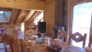 A cozy wooden dining area with a table set for four, surrounded by wooden chairs with heart-shaped cutouts. A television is mounted on the wall, and a fireplace is visible in the background.