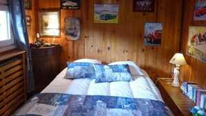 Una habitación con paneles de madera, una cama, carteles, una lámpara y una pequeña mesa con libros.