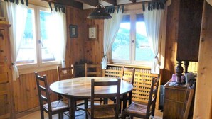 Un comedor de madera con una mesa redonda y seis sillas, ventanas amplias con cortinas y vistas al exterior.