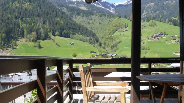 Una terraza de madera con banco y mesas, con vista a un paisaje montañoso con casas.