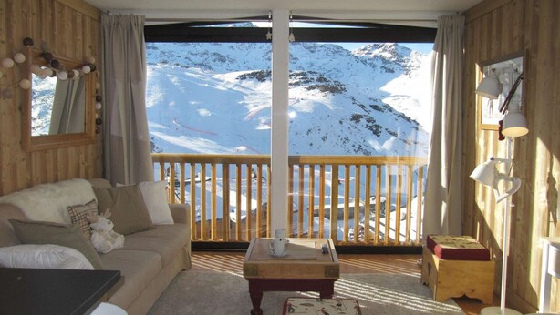A cozy room with a wooden interior, a large window offering a snowy mountain view, a wooden coffee table, and a sofa with a plush blanket.