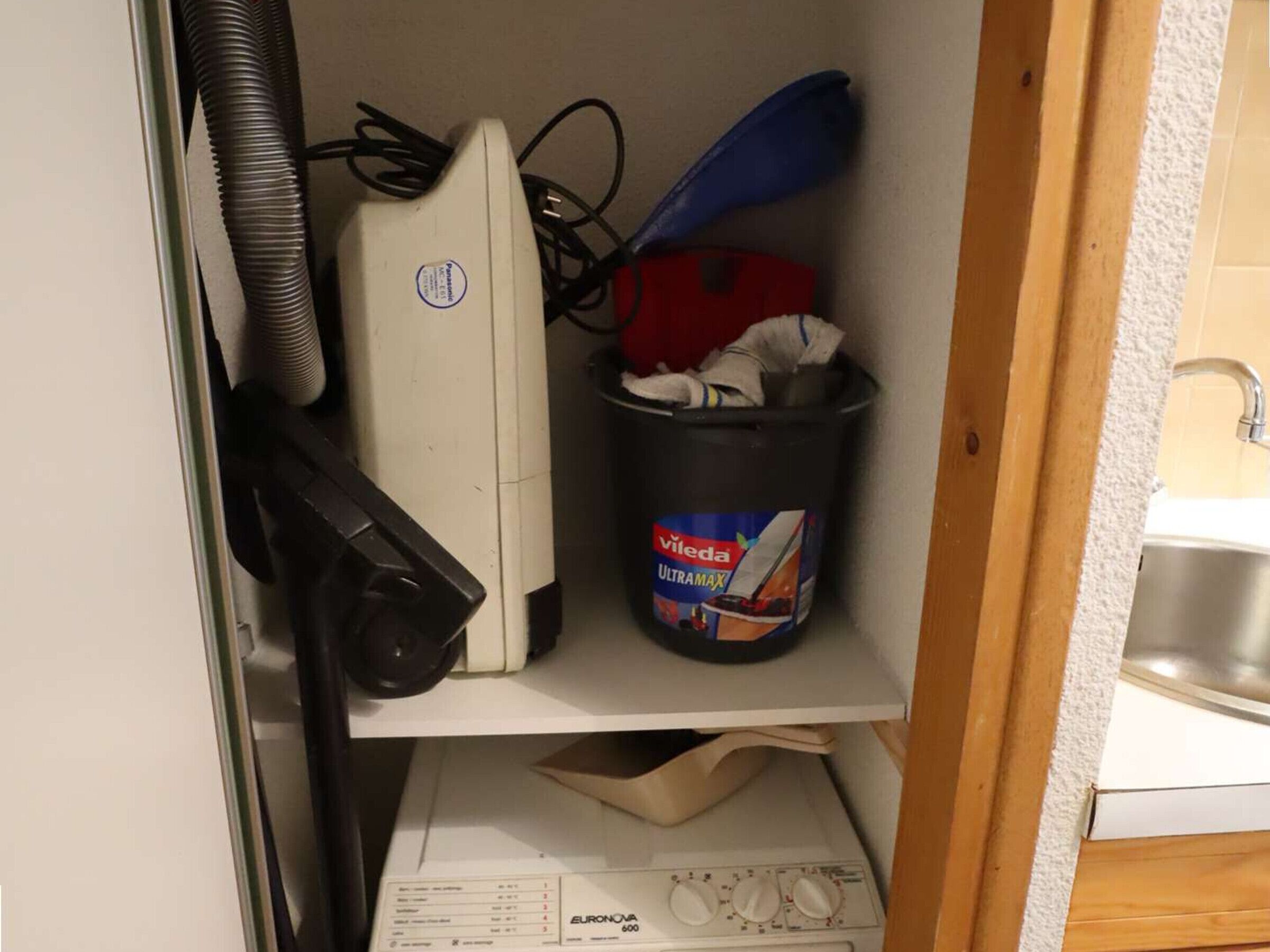 Een klein keukentje met een wit apparaat, een prullenbak met een label en een spoelbak.