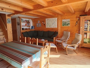 Interior de una cabaña de madera con una cama, un sofá, dos sillones, una mesa y una estantería.