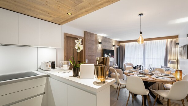 A modern kitchen with white cabinets, a central island with a sink, and a dining area with a wooden table and chairs. - Beautiful 76m² apartment in Courchevel, near slopes, 3 bedrooms, parking (Courchevel 1550)