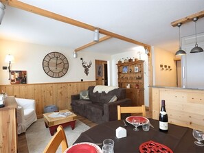Uma sala de estar aconchegante com teto de madeira, um relógio de parede redondo, uma decoração de cabeça de veado, e uma área de jantar com mesa posta para dois.
