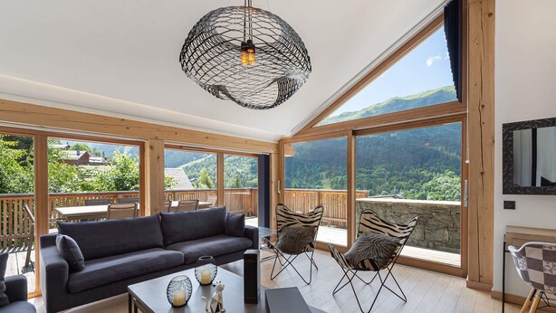 A spacious living room with a large sofa, a coffee table, and a TV mounted on the wall. There is a balcony with wooden railings and a view of the mountains. - Modern chalet in Méribel with sauna and jacuzzi, 6 bedrooms (Méribel)