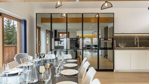 A dining area with a long table set for a meal, a kitchen in the background, and a view of the outdoors. - Modern chalet in Méribel with sauna and jacuzzi, 6 bedrooms (Méribel)