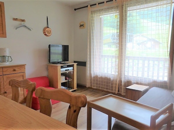 Ett mysigt vardagsrum med ett TV-bord i trä, en röd soffa, ett matbord och en utsikt genom ett fönster med en vit gardin.
