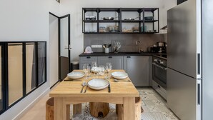 A modern kitchen with a wooden dining table set for four, a stainless steel refrigerator, and a black shelving unit above the counter.