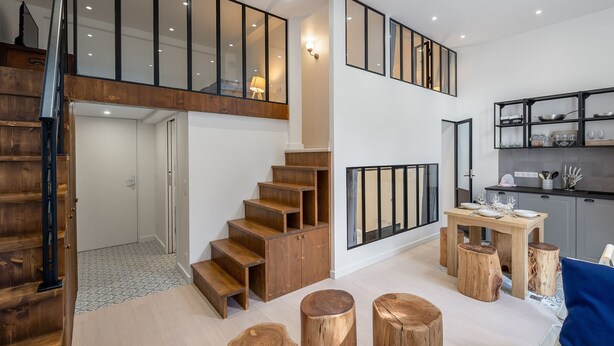 A modern interior with a wooden staircase, a dining area with stools, and a kitchen with open shelving.